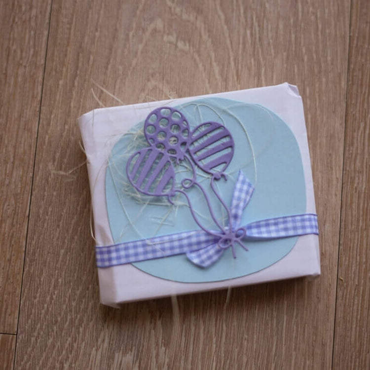 Ein kleines Geschenk, verpackt in weißem Papier mit blauer Schleife und verziert mit der Stanzschablone Drei Luftballons mit Muster von Stanzenshop.de, perfekt zum Basteln Geburtstag, liegt auf einer Holzfläche.