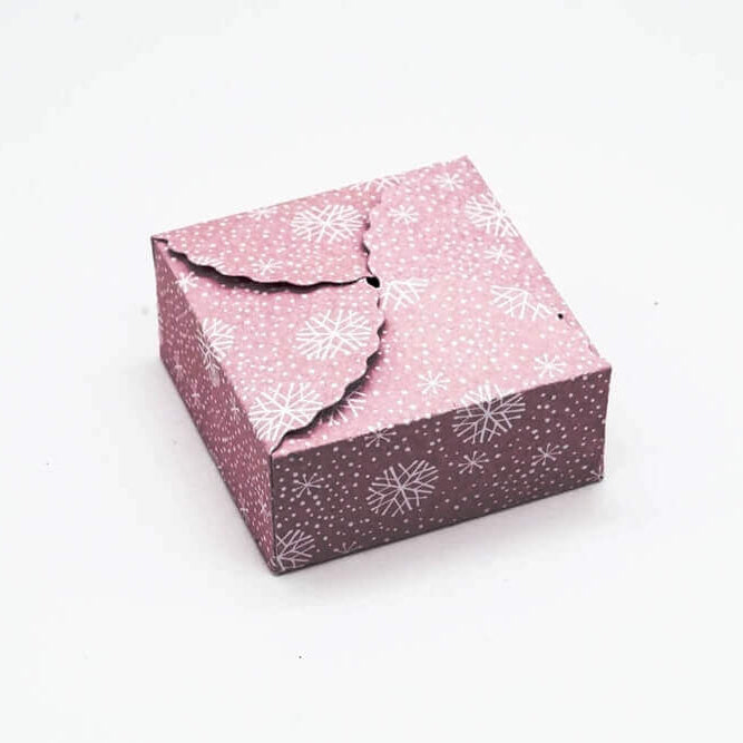 Ein kleines, quadratisches Stanzschablone Geschenkkästchen von Stanzenshop.de mit rosa Hintergrund und weißen Schneeflocken, ideal zum Basteln oder Scrapbooking, wird geschlossen und auf einer schlichten weißen Fläche ausgestellt.