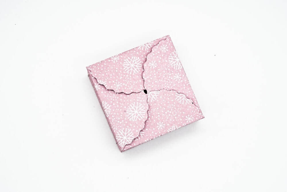 Die Stanzschablone Geschenkkästchen von Stanzenshop.de zeigt einen geschlossenen rosa Umschlag mit Schneeflockenmuster und Wellenkanten, perfekt zum Basteln oder Scrapbooking, abgebildet auf einem weißen Hintergrund.