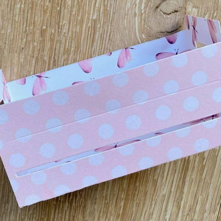 Eine rosafarbene rechteckige Pappschachtel mit weißen Tupfen und Schmetterlingsmuster im Inneren, hergestellt mit der Stanzschablone: Großer Karton von Stanzenshop.de, perfekt als Geschenkbox Hochzeit und für Bastelprojekte, abgebildet auf einer Holzunterlage.