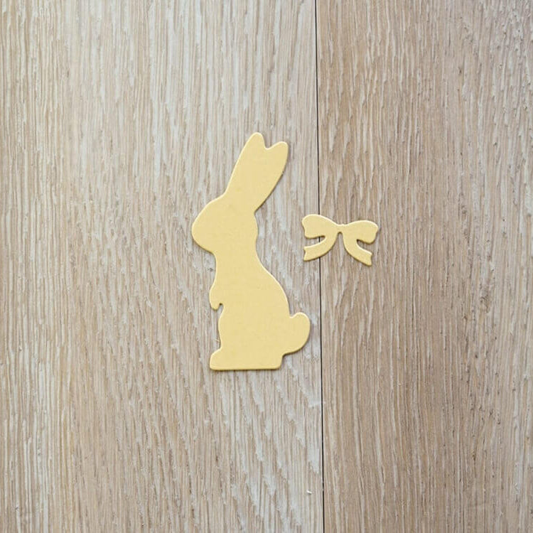 Ein gelber Papierausschnitt eines sitzenden Hasen mit einer Schleife, hergestellt mit der "Stanzschablone: Hase mit einer Fliege" von Stanzenshop.de, wird auf hellem Holz präsentiert - eine perfekte Idee fürs Basteln zu Ostern.