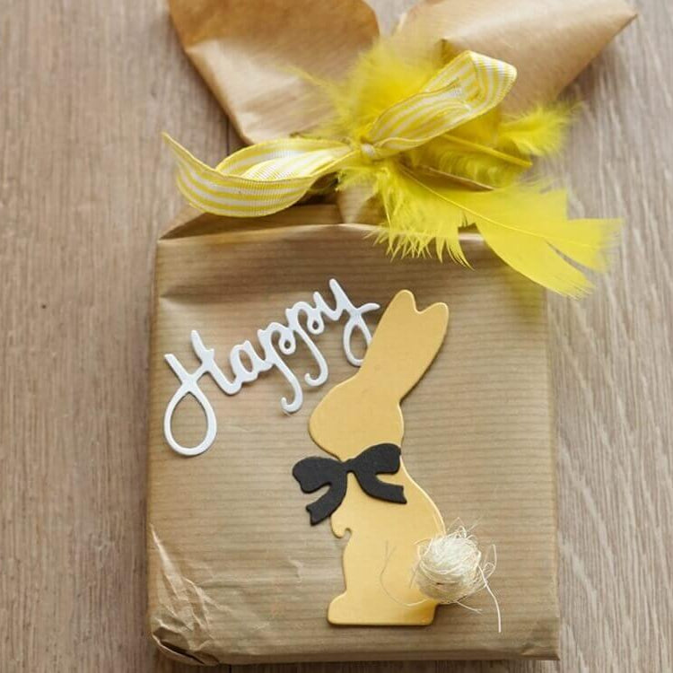 Ein in braunes Papier eingepacktes Geschenk mit gelben Federn, einem gelben Papierhasen aus Stanzschablone: Hase mit einer Fliege von Stanzenshop.de, einer kleinen Schleife und "Happy" oben drauf - ideal zum Basteln zu Ostern.