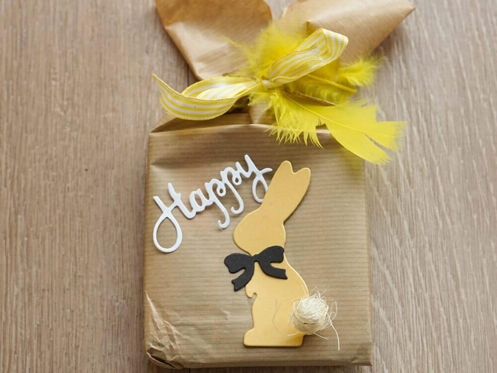 Ein in braunes Papier eingepacktes Geschenk mit gelben Federn, einem gelben Papierhasen aus Stanzschablone: Hase mit einer Fliege von Stanzenshop.de, einer kleinen Schleife und "Happy" oben drauf - ideal zum Basteln zu Ostern.