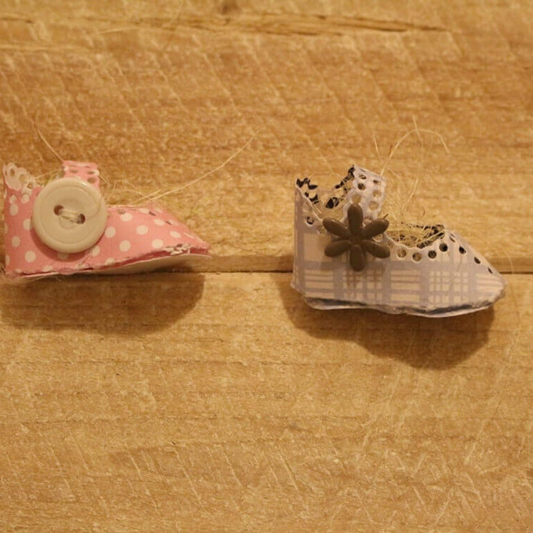 Zwei kleine handgefertigte Babyschuh-Dekorationen, hergestellt mit der "Stanzschablone Kinderschuh für ein Baby" von Stanzenshop.de - eine rosafarbene mit einem weißen Knopf und eine blaue mit einem Blumenknopf - sind ideal für Scrapbooking-Projekte.