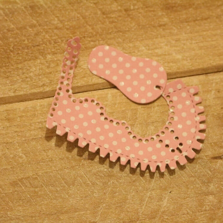 Zwei Stücke rosafarbenes, gepunktetes Papier, zugeschnitten mit der "Stanzschablone Kinderschuh für ein Baby" von Stanzenshop.de, werden dekorativ geformt und auf eine Holzunterlage gelegt.