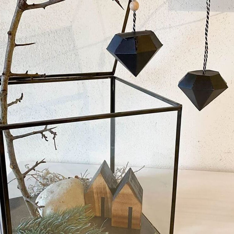 In einem Glasterrarium befinden sich ein Zweig, Moos, Holzhäuser und ein weißer Stein; darüber hängen zwei schwarze Stanzenshop.de "Stanzschablone Kleine Diamantschachtel", die für geometrischen Charme sorgen.