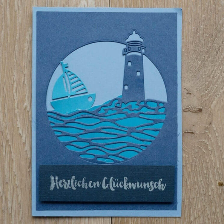 Die Stanzschablone Leuchtturm und Meer in rundem Rahmen von Stanzenshop.de schneidet eine blau-weiße Segelboot- und Leuchtturmszene mit "Herzlichen Glückwunsch" aus, perfekt für Basteln oder Scrapbooking-Projekte.