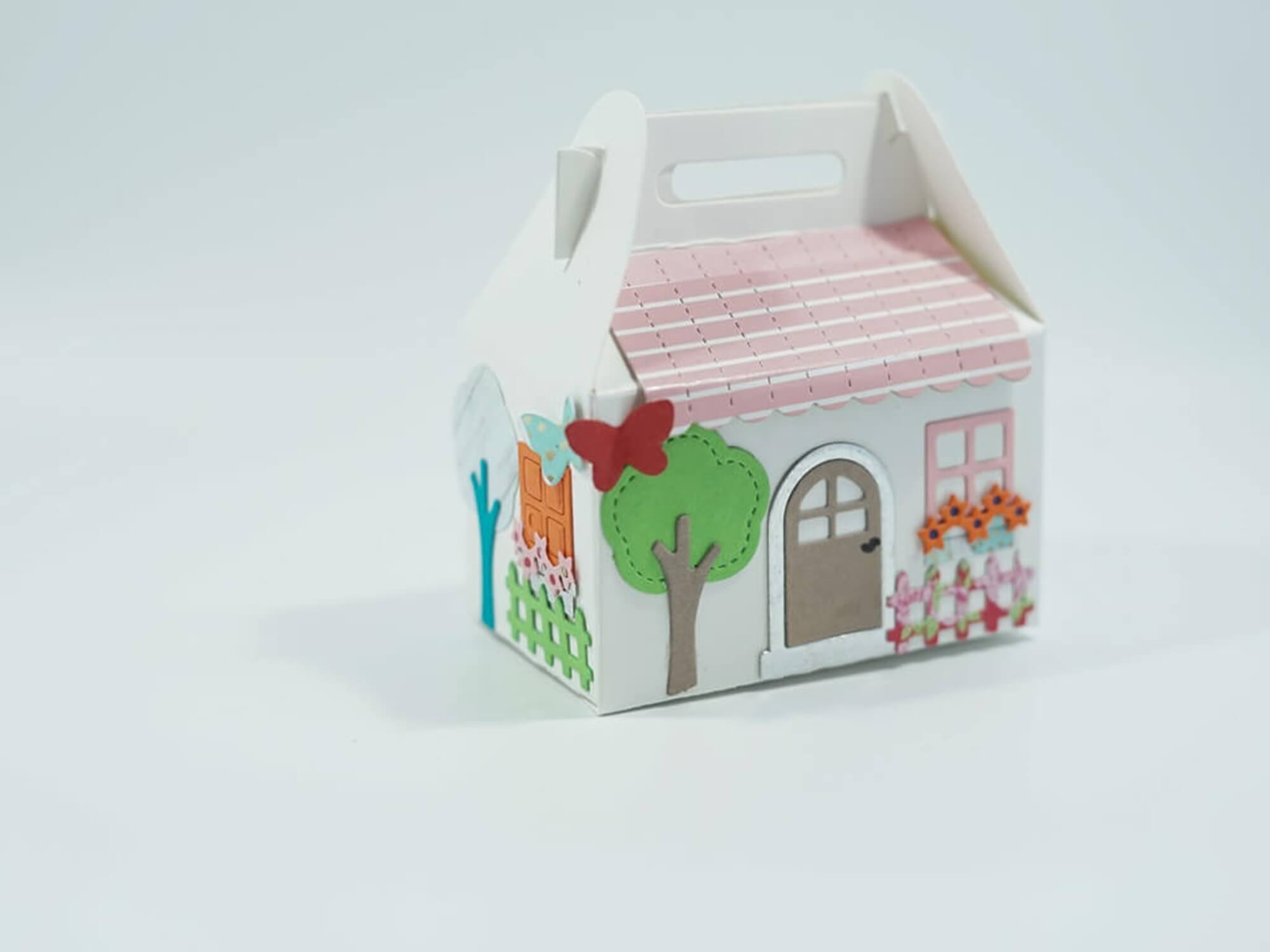 Charmante kleine Geschenkbox aus Pappe in Form eines Hauses mit rosa Dach, Bäumen, Schmetterling und Blumen - perfekt zum Osterbasteln mit der "Stanzschablone: Osterset" von Stanzenshop.de.