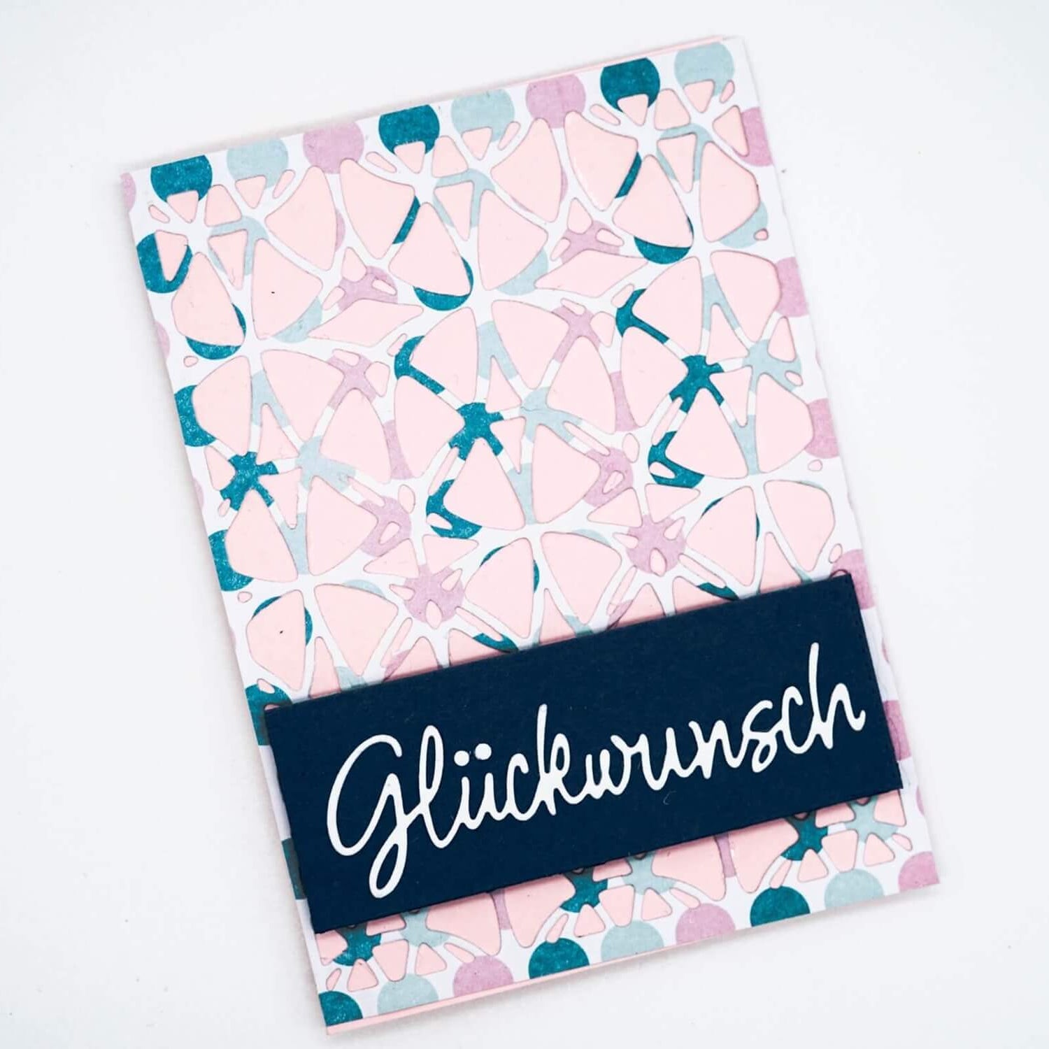 Die "Stanzschablone: Platte mit Kreismuster" von Stanzenshop.de kreiert abstrakte rosa und tealfarbene Kreismuster, ideal für Grußkarten mit "Glückwunsch" in weißer Schrift auf einem dunkelblauen Rechteck - perfekt für die Kartenherstellung.
