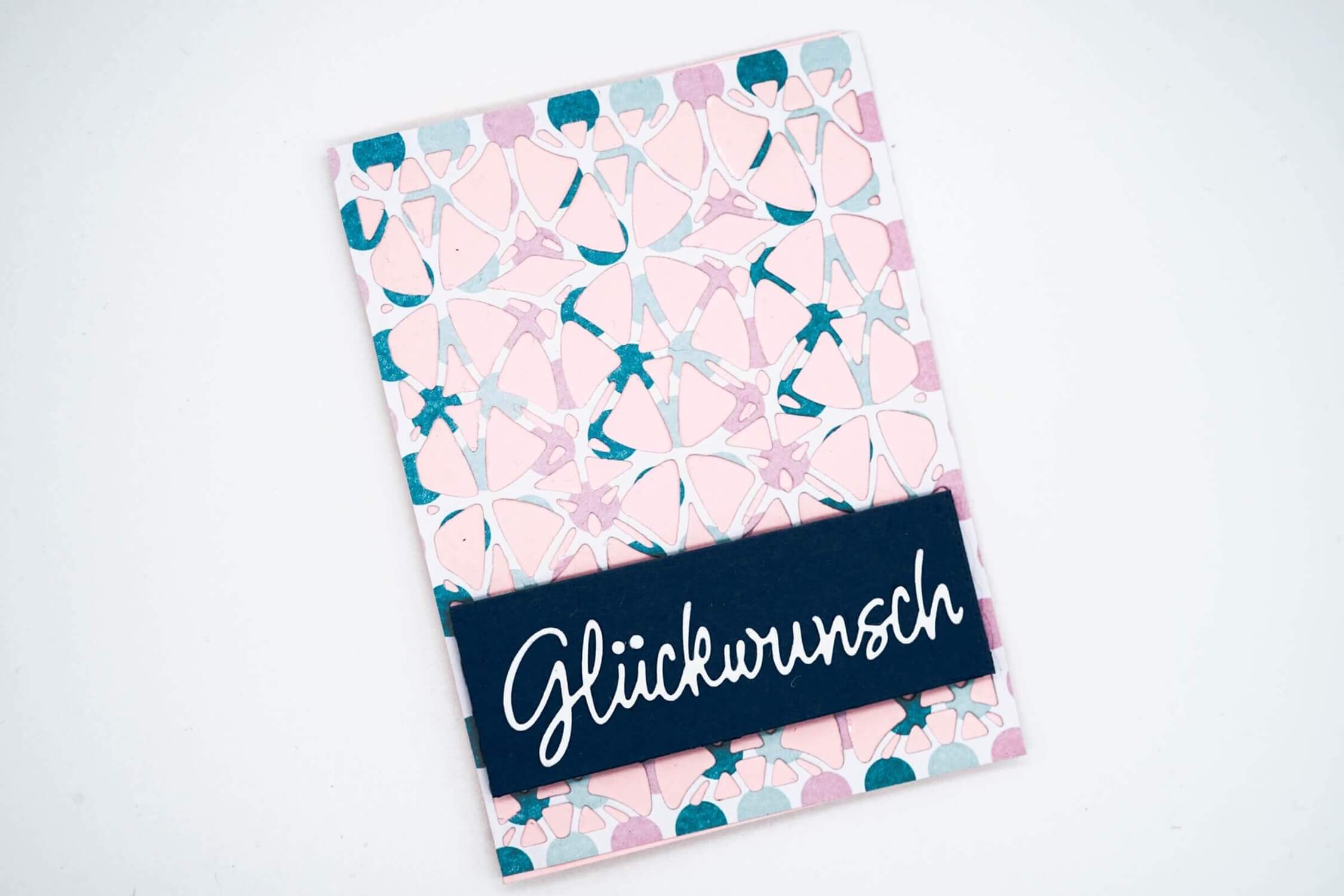 Die "Stanzschablone: Platte mit Kreismuster" von Stanzenshop.de kreiert abstrakte rosa und tealfarbene Kreismuster, ideal für Grußkarten mit "Glückwunsch" in weißer Schrift auf einem dunkelblauen Rechteck - perfekt für die Kartenherstellung.