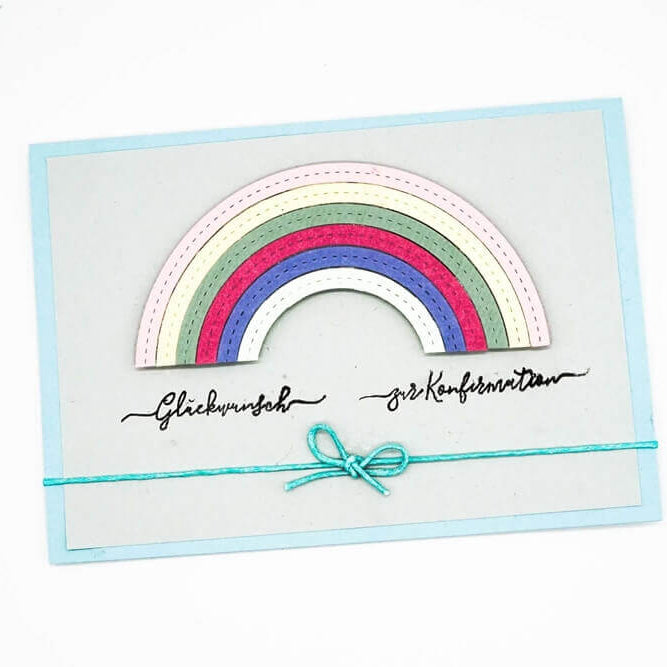 Eine Grußkarte mit einem geschichteten Regenbogendesign mit der Stanzschablone Regenbogen von Stanzenshop.de, handgeschriebenem deutschen Text und einer blauen Schleife - ein inspirierendes Bastelergebnis, kompatibel mit gängigen Stanzmaschinen.