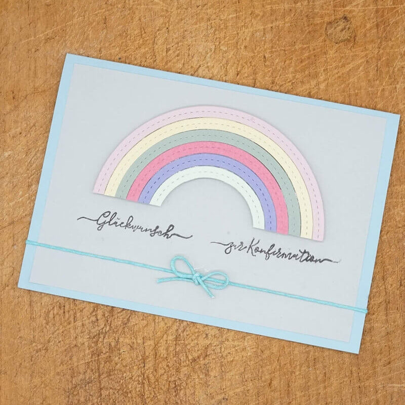 Die pastellfarbene Regenbogenkarte aus der Stanzschablone Regenbogen von Stanzenshop.de mit kursiver Schrift und blauer Schnurschleife ruht auf Holz - perfekt, um Ihr Bastelergebnis zu präsentieren.