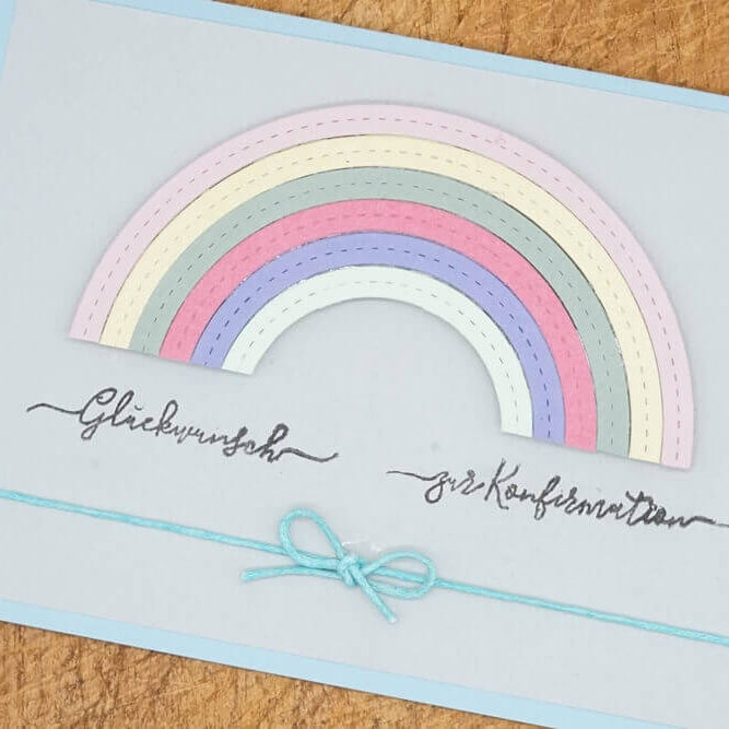 Eine handgefertigte Karte von Stanzenshop.de mit der Stanzschablone Regenbogen in Pastelltönen, deutschem Text und einer blauen Schleife, dargestellt auf einer Holzfläche.