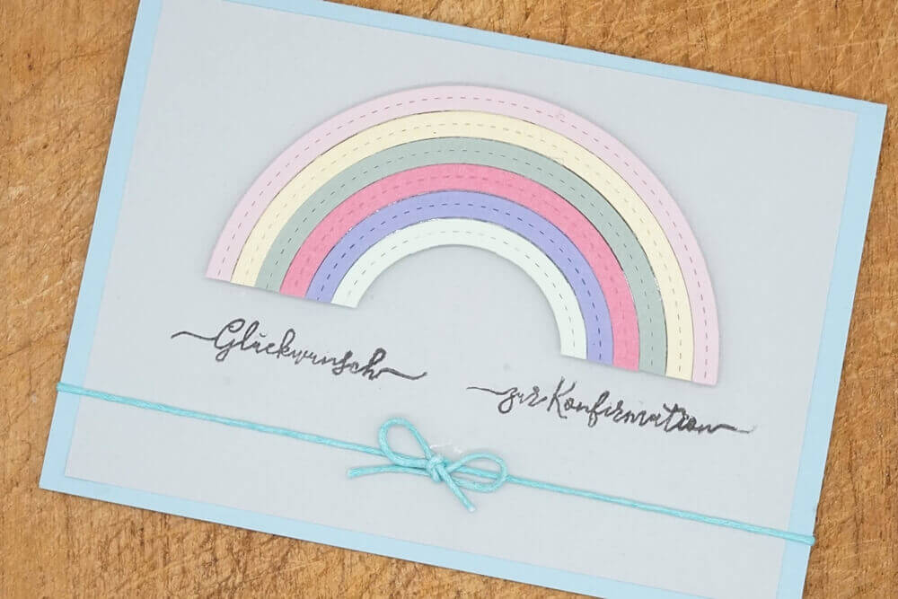 Eine handgefertigte Karte von Stanzenshop.de mit der Stanzschablone Regenbogen in Pastelltönen, deutschem Text und einer blauen Schleife, dargestellt auf einer Holzfläche.