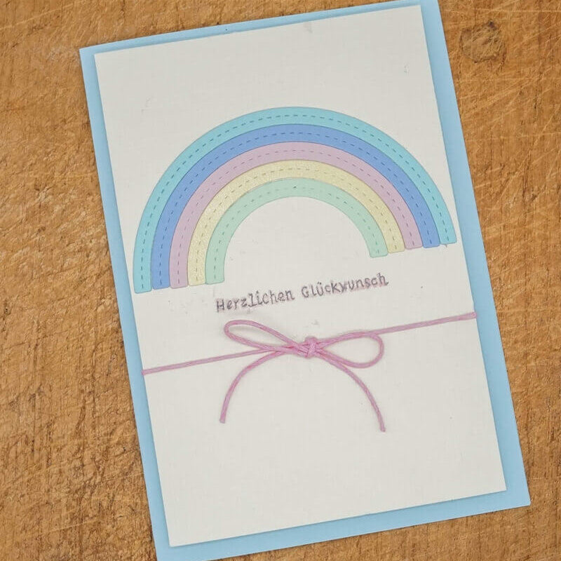 Eine Grußkarte mit einem pastellfarbenen Regenbogen aus der Stanzschablone Regenbogen von Stanzenshop.de, mit "Herzlichen Glückwunsch" auf Deutsch und einem rosafarbenen Streichbogen, ausgestellt auf Holz für ein inspirierendes Bastelergebnis.