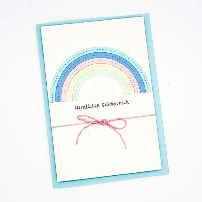 Eine Grußkarte mit der Stanzschablone Regenbogen von Stanzenshop.de zeigt ein pastellfarbenes Regenbogendesign, den Schriftzug "Herzlichen Glückwunsch" und eine rosa Schleife auf hellblauem Hintergrund.