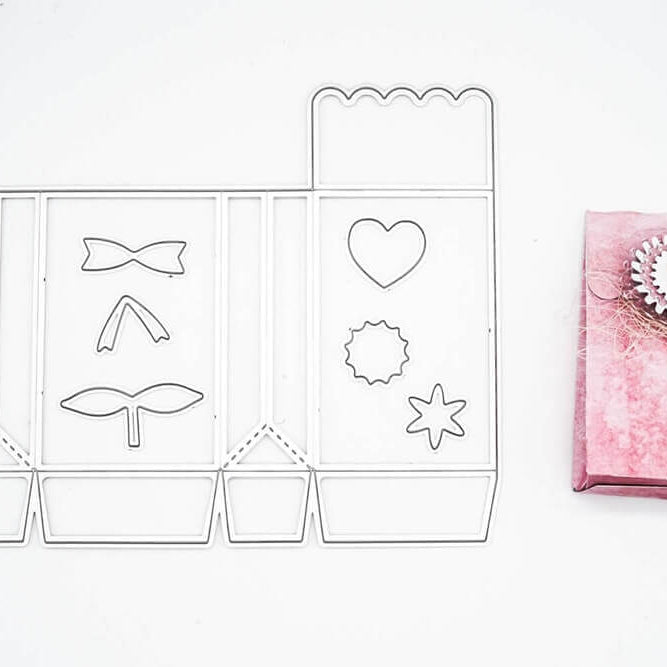 Die Stanzschablone Schachtel mit Verzierungen von Stanzenshop.de liegt neben einer rosafarbenen Geschenkbox mit kreisförmiger Verzierung - ideal zum Schachtel basteln oder zum Basteln von einzigartigen Gastgeschenken.