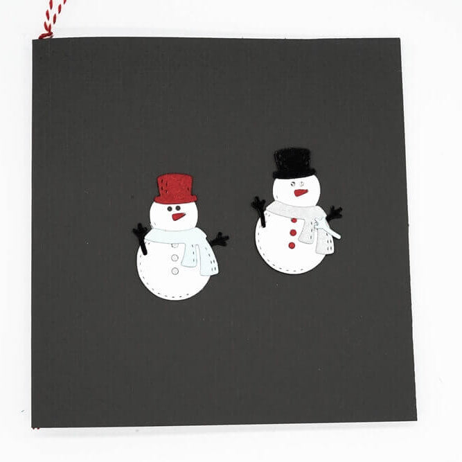 Eine schwarze Karte mit zwei Schneemännern - einer mit roter Mütze und Schal, einer mit schwarzer Mütze und weißem Schal - hergestellt mit der "Stanzschablone: Schneemann mit Schal und Zylinder" von Stanzenshop.de, perfekt für winterliche Bastel- oder weihnachtliche Stanzprojekte.