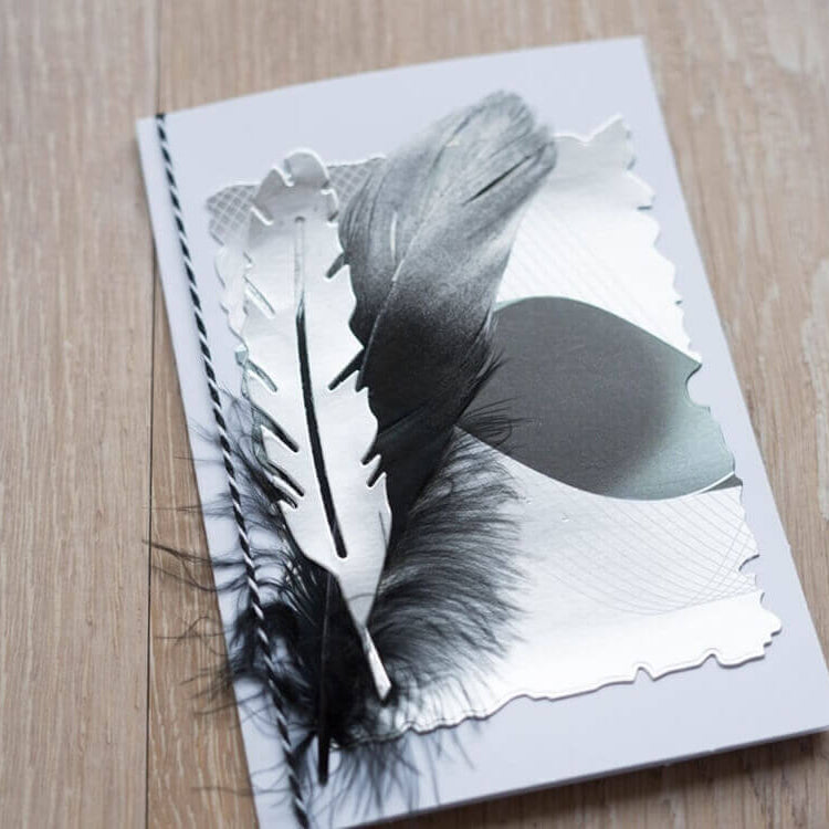 Eine handgemachte Scrapbooking-Karte mit strukturiertem Papier und einer schwarzen Feder, hergestellt mit der "Stanzschablone: Sechs gewellte Rahmen in verschiedenen Größen" von Stanzenshop.de, die auf einer Holzoberfläche präsentiert wird.