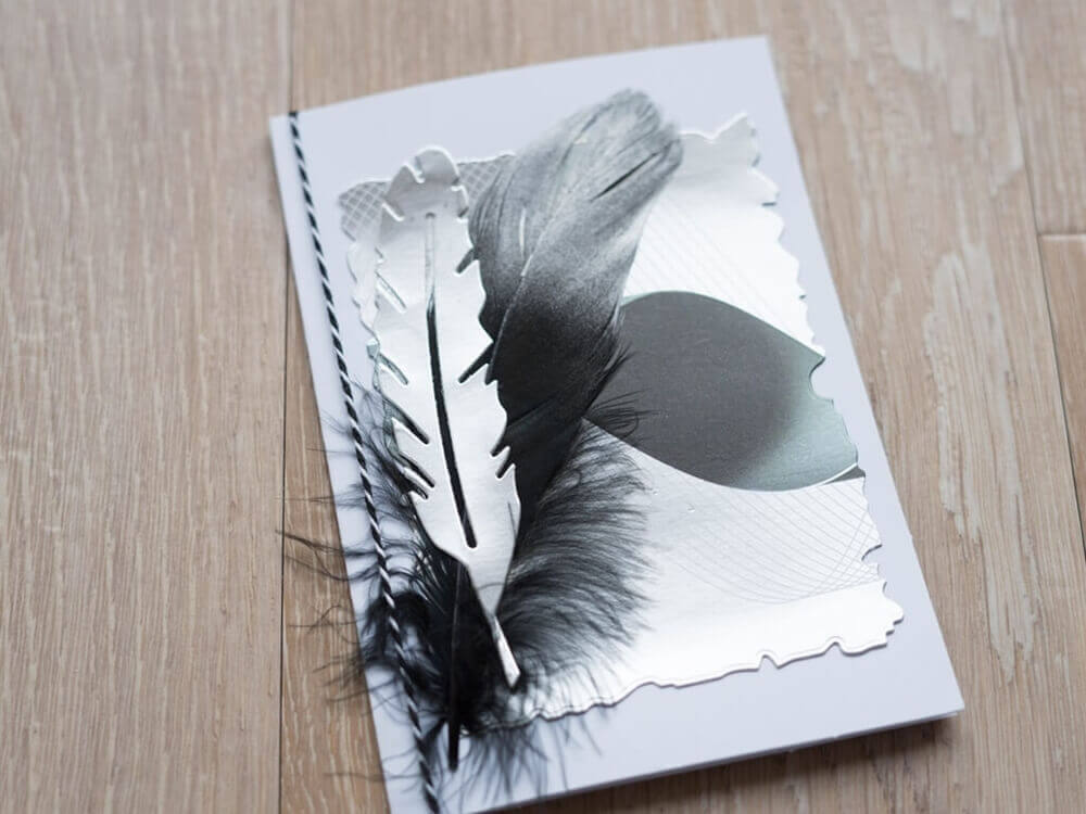 Eine handgemachte Scrapbooking-Karte mit strukturiertem Papier und einer schwarzen Feder, hergestellt mit der "Stanzschablone: Sechs gewellte Rahmen in verschiedenen Größen" von Stanzenshop.de, die auf einer Holzoberfläche präsentiert wird.