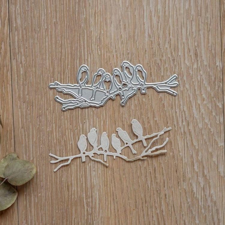 Die Stanzschablone "Sechs Vögel auf einem Ast" von Stanzenshop.de zeigt sechs Vögel auf einem Ast, dargestellt auf Holz mit getrockneten Blättern - ideal für Kartenherstellung oder Scrapbooking.