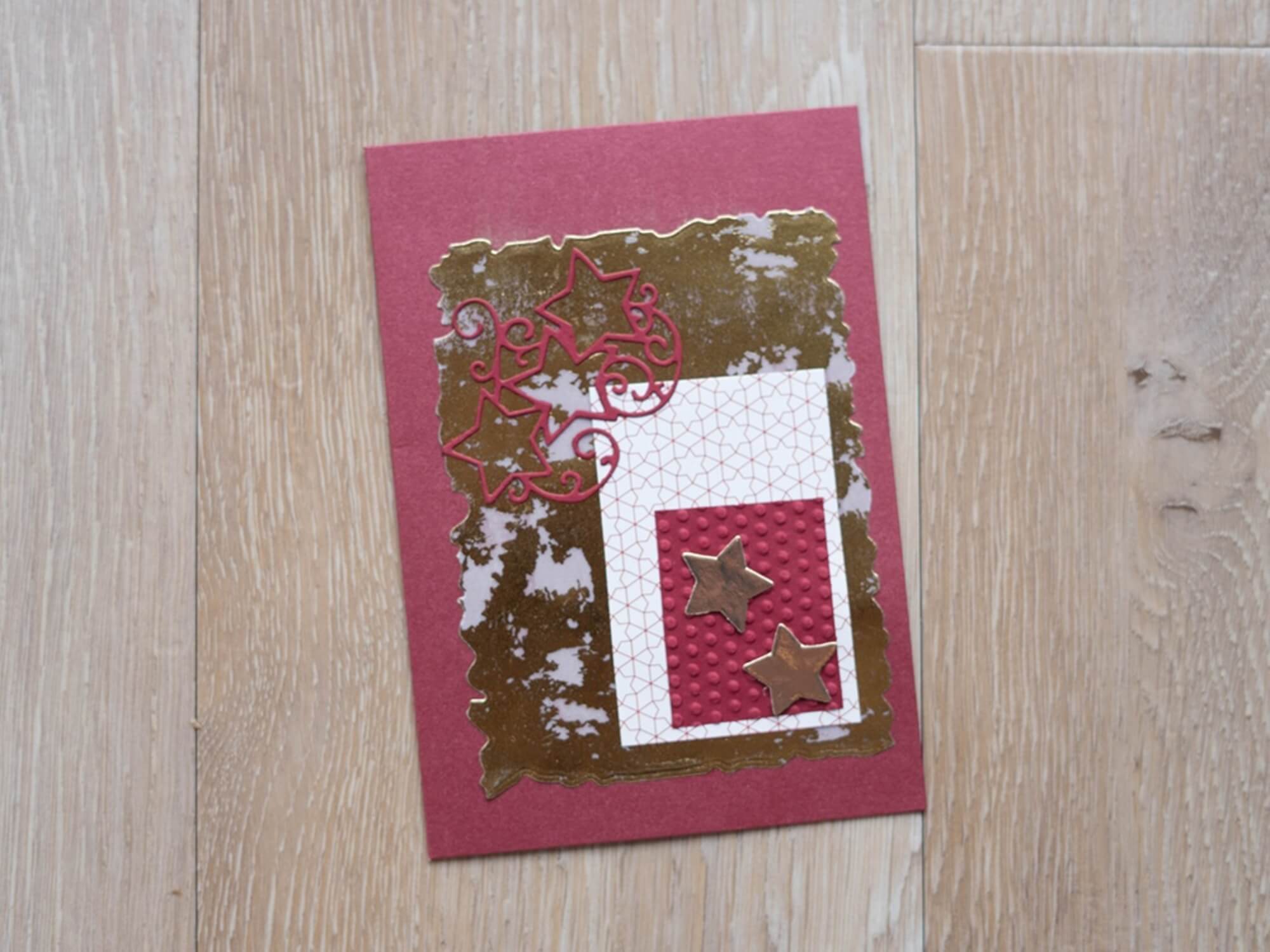 Eine handgefertigte Grußkarte von Stanzenshop.de, hergestellt mit der "Stanzschablone: Stern mit Rahmen", mit goldenen und rosafarbenen Sterndekorationen auf rotem, mit Goldfolie akzentuiertem und strukturiertem Papier, auf einer Holzoberfläche.