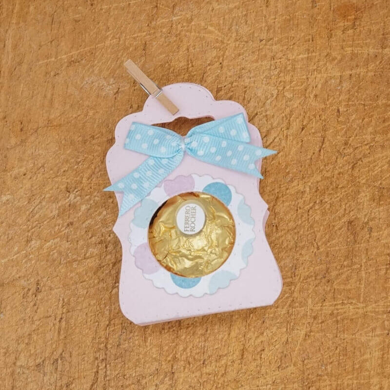Ein Ferrero Rocher auf einer pastellrosa und blauen Karte, hergestellt mit der Stanzschablone Tischkarte mit Kreis von Stanzenshop.de, verziert mit einer blauen Tupfenschleife - ideal für Scrapbooking oder Bastelset-Projekte.