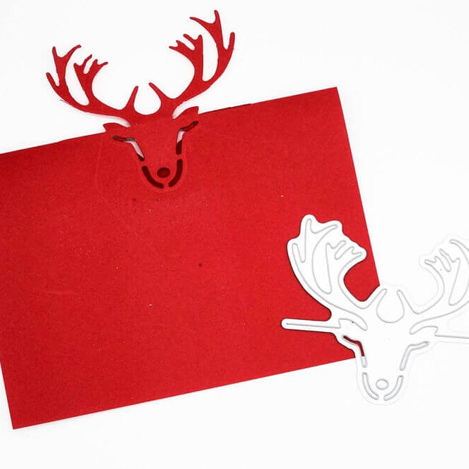 Eine rote Karte mit einem ausgeschnittenen Rentierkopf aus der Stanzschablone: Die Tischkarte Rentierkopf von Stanzenshop.de liegt neben einer weißen Rentierschablone aus Metall - ideal für Tischkarte Basteln oder Scrapbooking Schablonen Projekte.