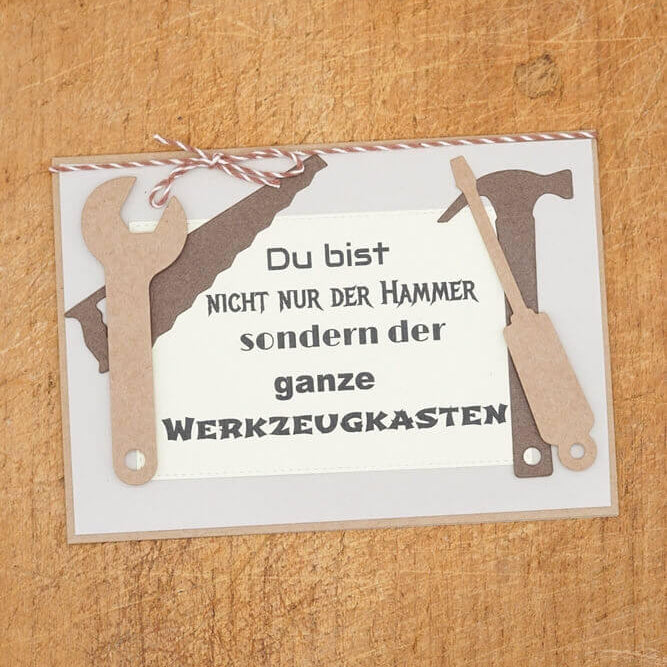 Eine Karte mit deutschem Text, verziert mit der "Stanzschablone Vier Werkzeuge" von Stanzenshop.de mit Ausschnitten eines Schraubenschlüssels, Hammers und einer Säge, ist mit brauner und weißer Schnur gebunden - ideal für Scrapbooking-Projekte.
