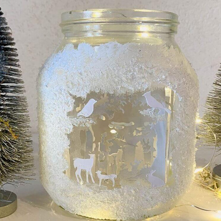 Ein mit Kunstschnee und Ausschnitten dekoriertes Glasgefäß aus der Stanzschablone Wald mit Tieren von Stanzenshop.de, platziert zwischen zwei verschneiten, mit Lichterketten geschmückten Kunstbäumchen.