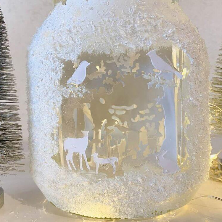 Ein Glasgefäß mit weißen Silhouetten von Rehen und Vögeln aus der Stanzschablone Wald mit Tieren von Stanzenshop.de, umgeben von Kunstschnee und Mini-Tannenbäumen - ein reizvolles Bastelergebnis für winterliches Scrapbooking.
