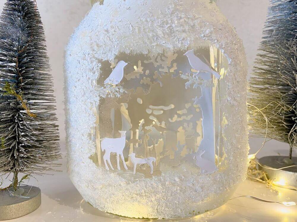 Ein Glasgefäß mit weißen Silhouetten von Rehen und Vögeln aus der Stanzschablone Wald mit Tieren von Stanzenshop.de, umgeben von Kunstschnee und Mini-Tannenbäumen - ein reizvolles Bastelergebnis für winterliches Scrapbooking.