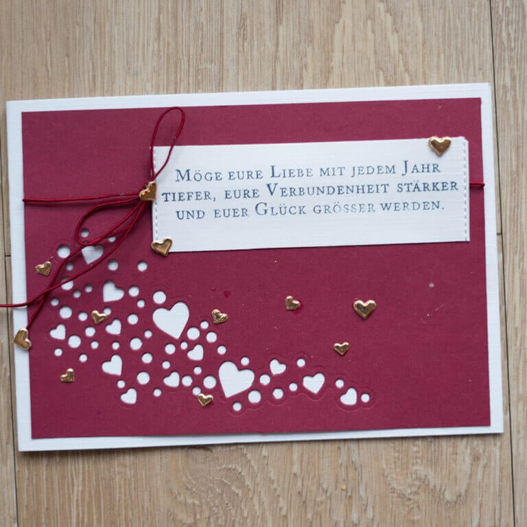 Die Stanzenschablone Welle aus Herzen von Stanzenshop.de kreiert burgunderfarbene handgefertigte Karten mit kleinen Herzausschnitten, goldenen Herzakzenten und einer deutschen Botschaft über die Liebe - ideal für Scrapbooking- oder Bastelprojekte.