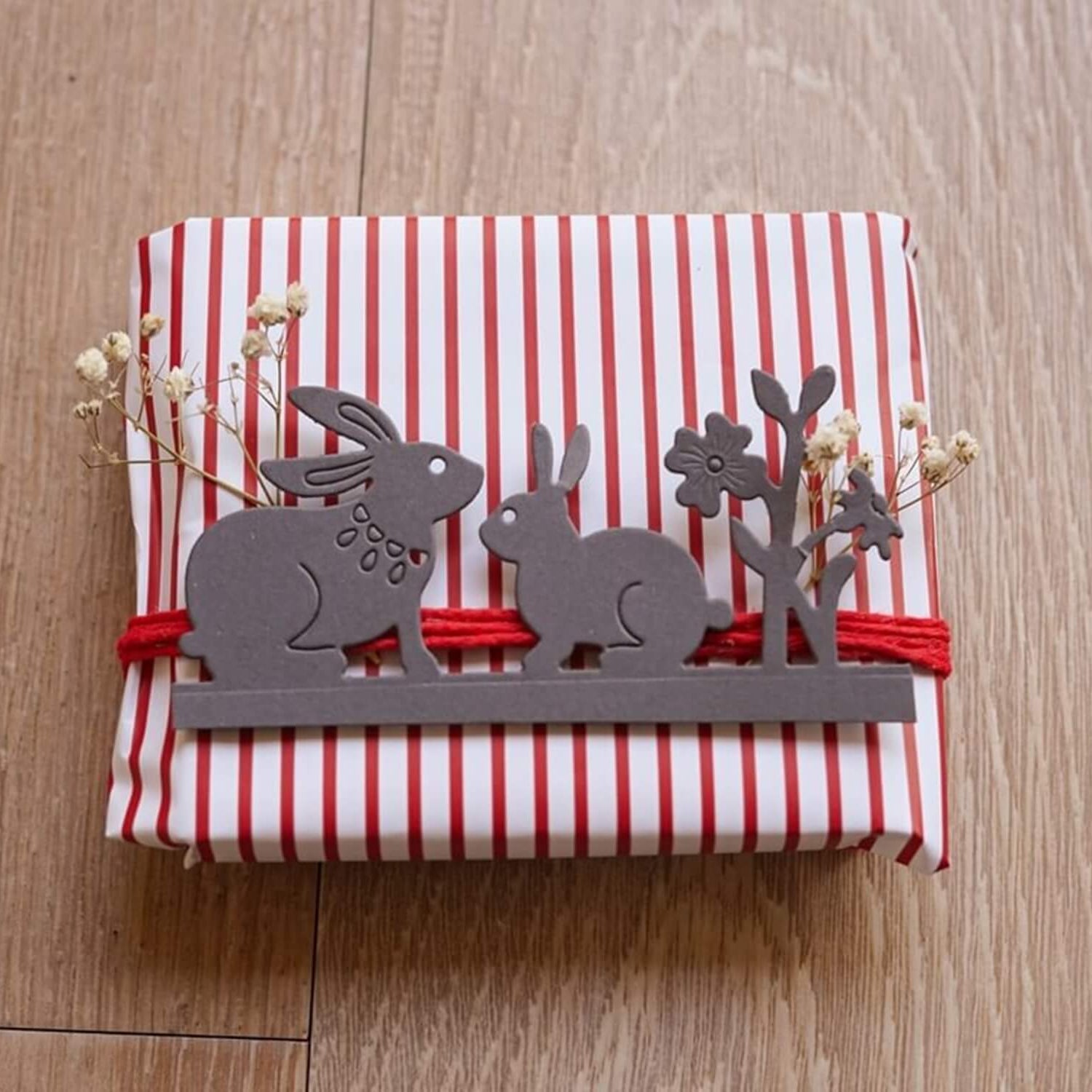 Ein in rot-weiß gestreiftes Papier verpacktes Geschenk, verziert mit zwei Hasen und einer Blume, die mit der Stanzschablone ausgeschnitten wurden: Zwei Hasen und eine Blume von Stanzenshop.de, dazu rote Schnur, kleine weiße Blumen und ein ausgeschnittener Baum. Ideal für Bastel-Fans.