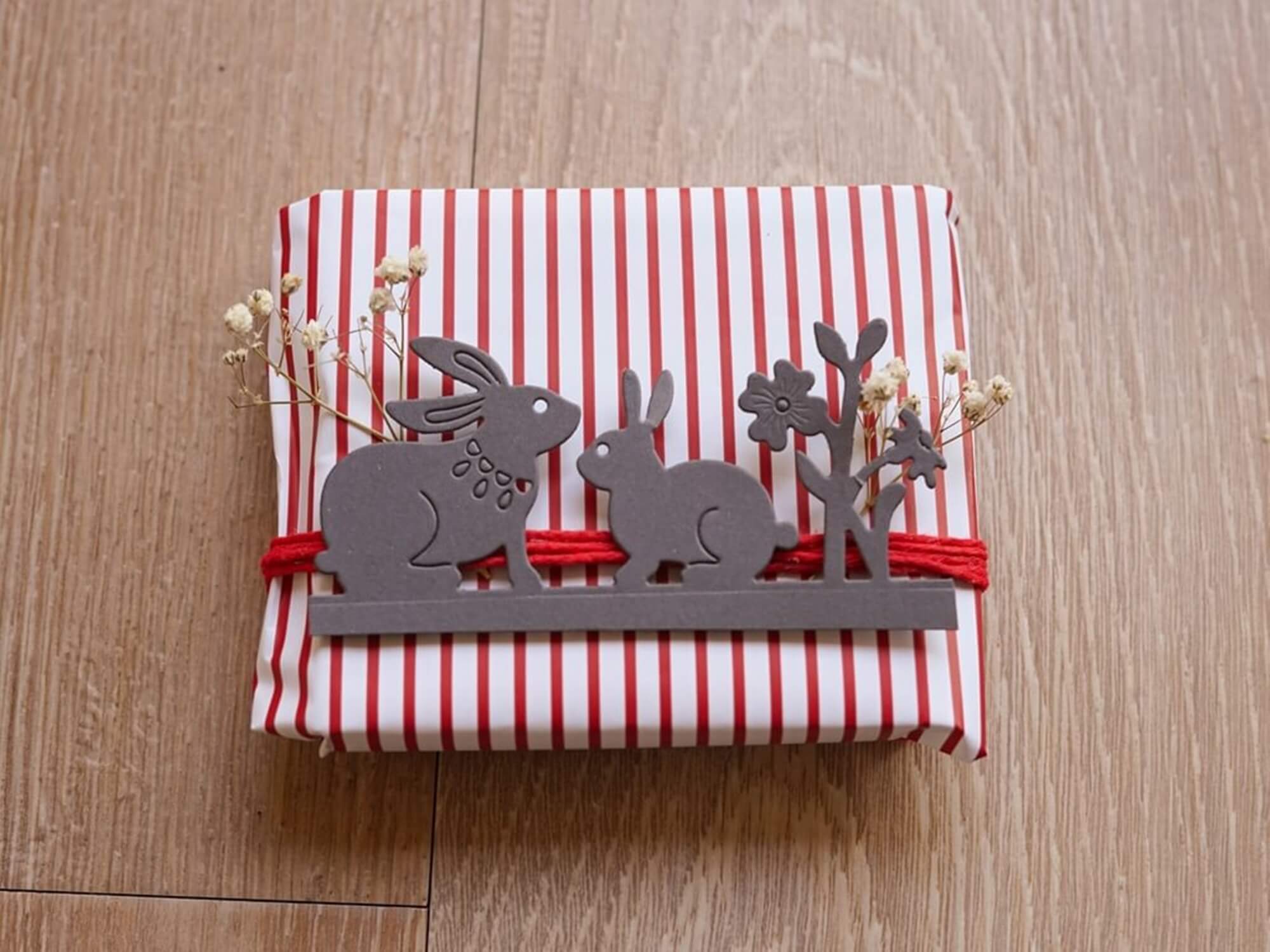 Ein in rot-weiß gestreiftes Papier verpacktes Geschenk, verziert mit zwei Hasen und einer Blume, die mit der Stanzschablone ausgeschnitten wurden: Zwei Hasen und eine Blume von Stanzenshop.de, dazu rote Schnur, kleine weiße Blumen und ein ausgeschnittener Baum. Ideal für Bastel-Fans.