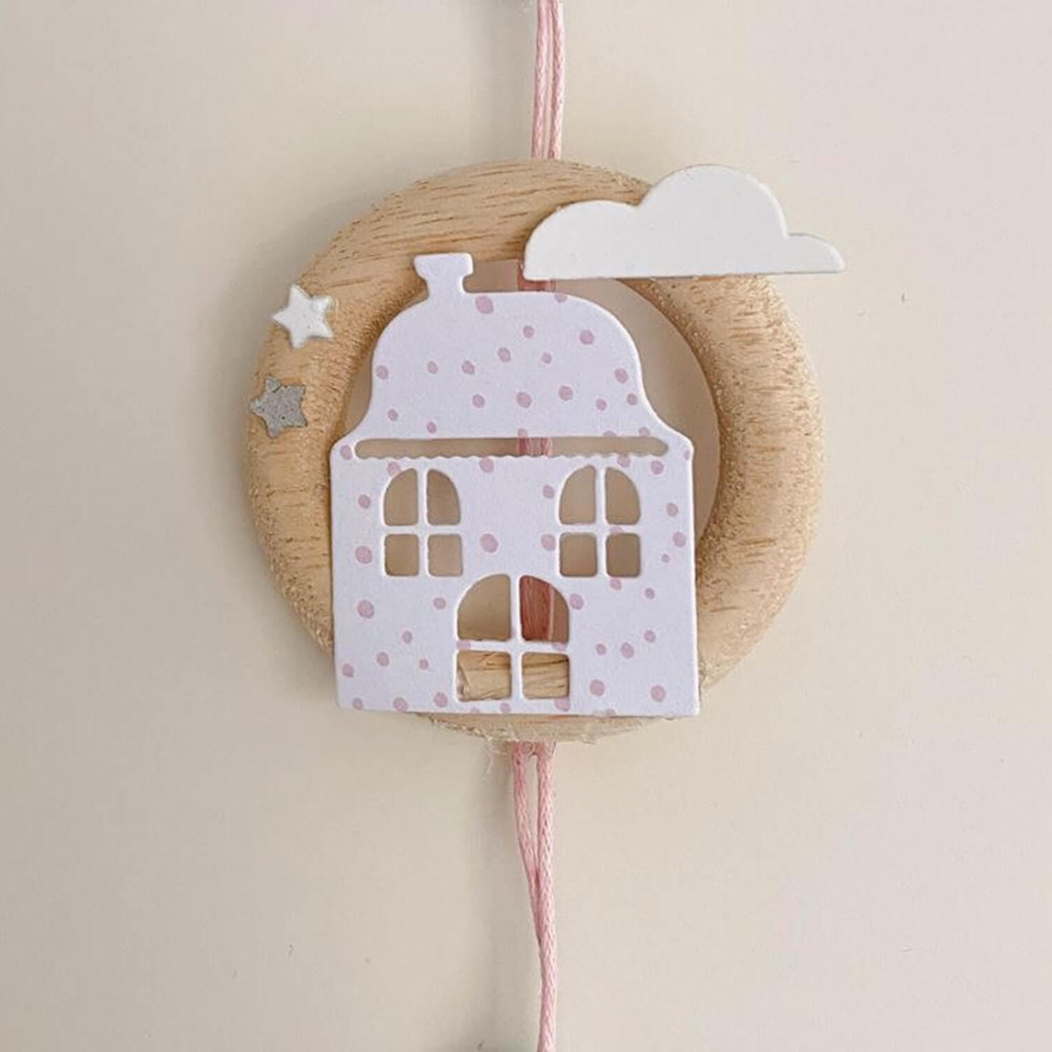 Ein rundes Holzornament mit rosa gepunktetem Haus, weißer Wolke und Sternen hängt an einer rosa Schnur - perfekt, um die "Stanzschablone: Zwei verschiedene Häuser" von Stanzenshop.de als Highlight vor hellen Hintergründen.
