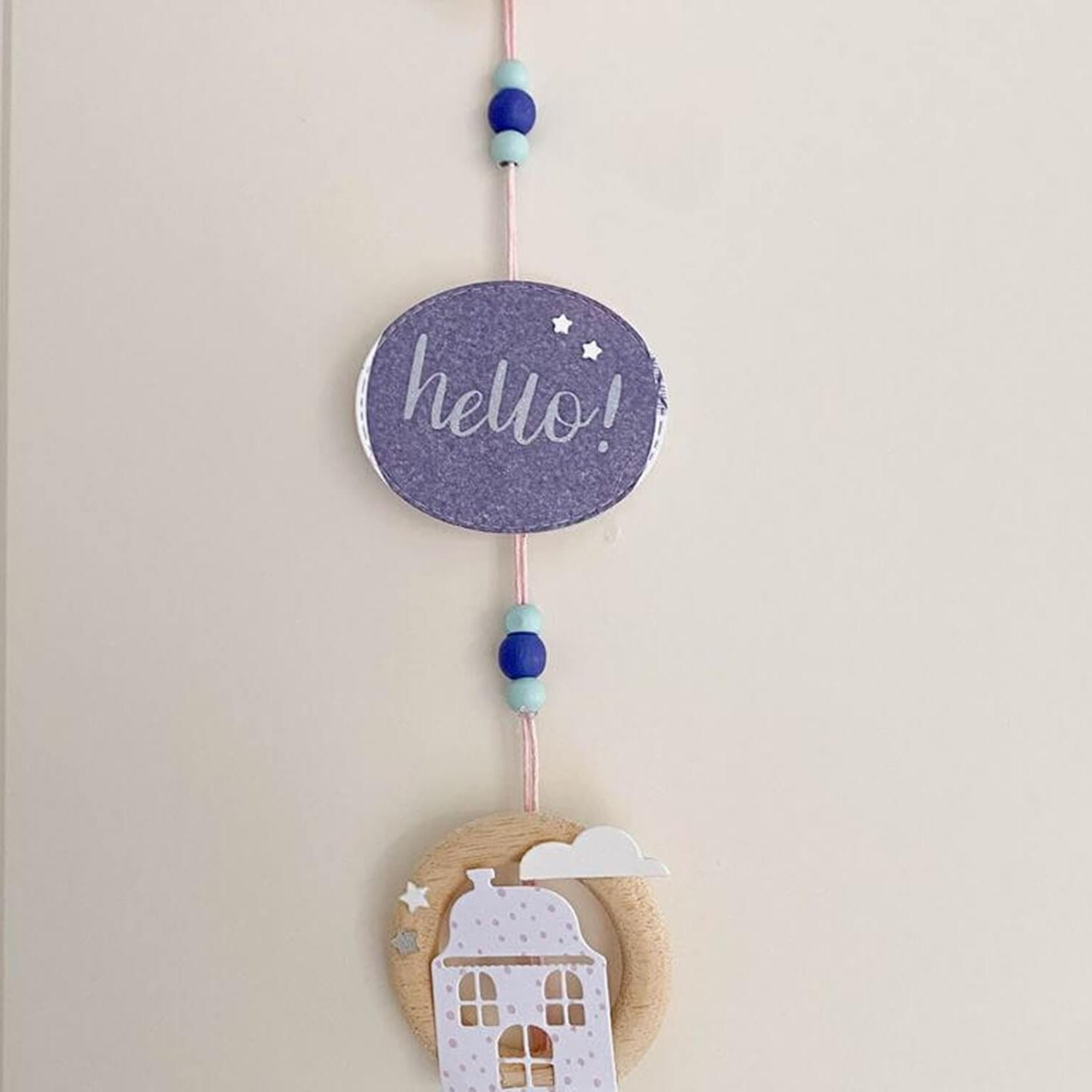 Eine handgefertigte Hängedekoration mit einem lila "Hallo!"-Schild, blauen Perlen und einem runden Holzteil mit weißem Hausmotiv - perfekt für Bastelprojekte mit der Stanzschablone: Zwei verschiedene Häuser von Stanzenshop.de.