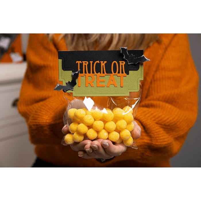 Eine Person in einem orangefarbenen Pullover hält die Sizzix 665951 - Thinlits Halloween Toppers von Sizzix, die eine durchsichtige Verpackung mit gelben Süßigkeitenkugeln, ein "Trick or Treat"-Etikett, dekorative schwarze Fledermäuse und einen 7,13x1-Aufkleber enthalten.