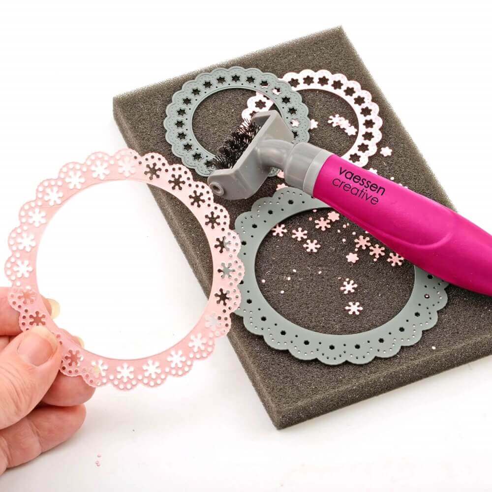 Eine Hand hält einen rosa, blumengemusterten Papierausschnitt neben dem Vaessen Creative Die brush & foam pad von Vaessen Creative.