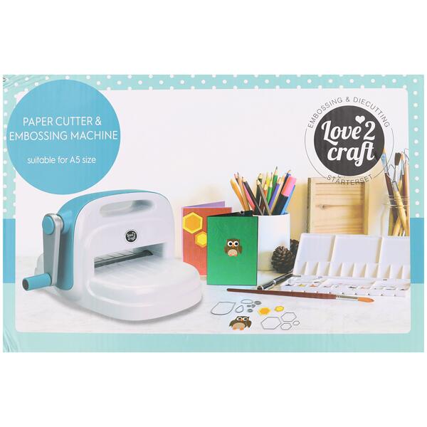 Eine Love 2 Craft Papierschneide- und Prägemaschine steht auf einem Schreibtisch mit Bastelmaterial, darunter Papier, Farbstifte und fertige Bastelprojekte.