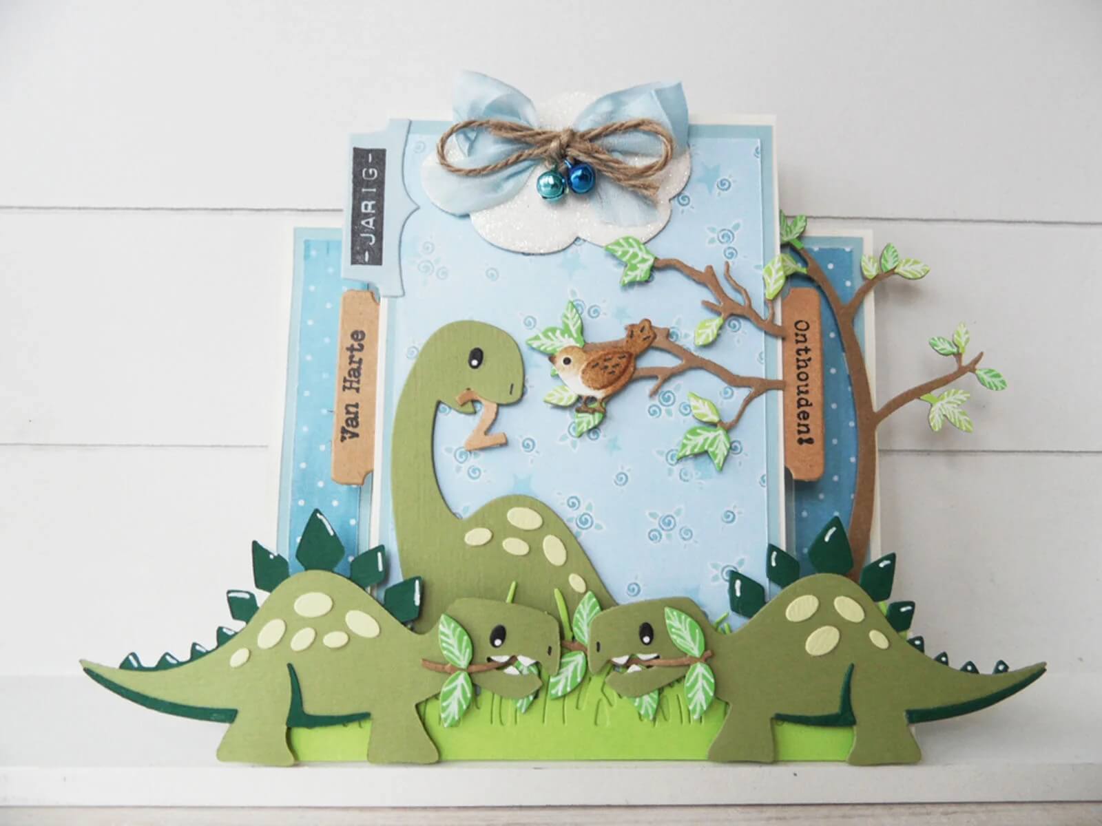 Handgefertigte Karte mit Marianne Design - Collectables Präge- und Stanzschablone Eline's Dinosaurier von Marianne Design, mit Cartoon-Dinosauriern, Blättern, Baum, Vogel, Wolke mit Schleife und Glöckchen; inklusive holländischen Grußanhängern. Ideal für Scrapbooking und Stanzmaschinen.