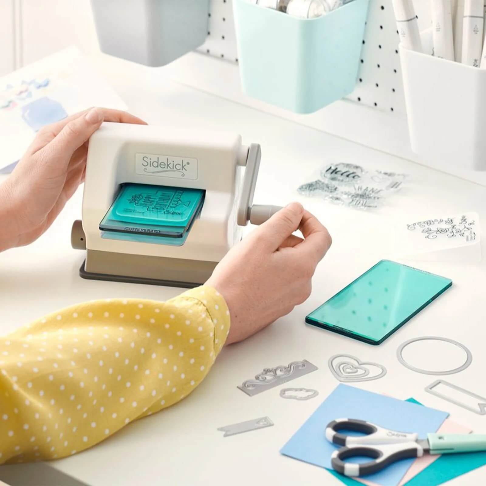 Ein Bastler verwendet das Sizzix 661770 - Sidekick Starter-Kit (Weiß & Grau) von Sizzix an einem Basteltisch mit Metallstempeln, Klarsichtstempeln, Scheren und verschiedenen Bastelsystem-Zubehörteilen.