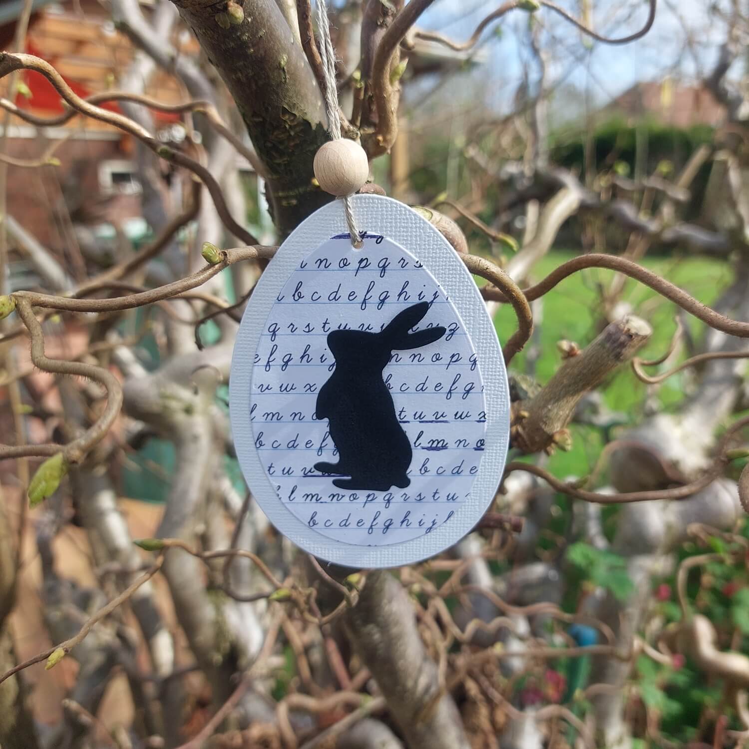 Ein ovales Ornament mit einer schwarzen Kaninchensilhouette, hergestellt mit der Stanzschablone: Drei verschiedene Kaninchen von Stanzenshop.de und kursiven Buchstaben hergestellt wurde, hängt an einem Baum im Freien - perfekt für Basteln zu Ostern und um Ihrer Osterdekoration handgemachten Charme zu verleihen.