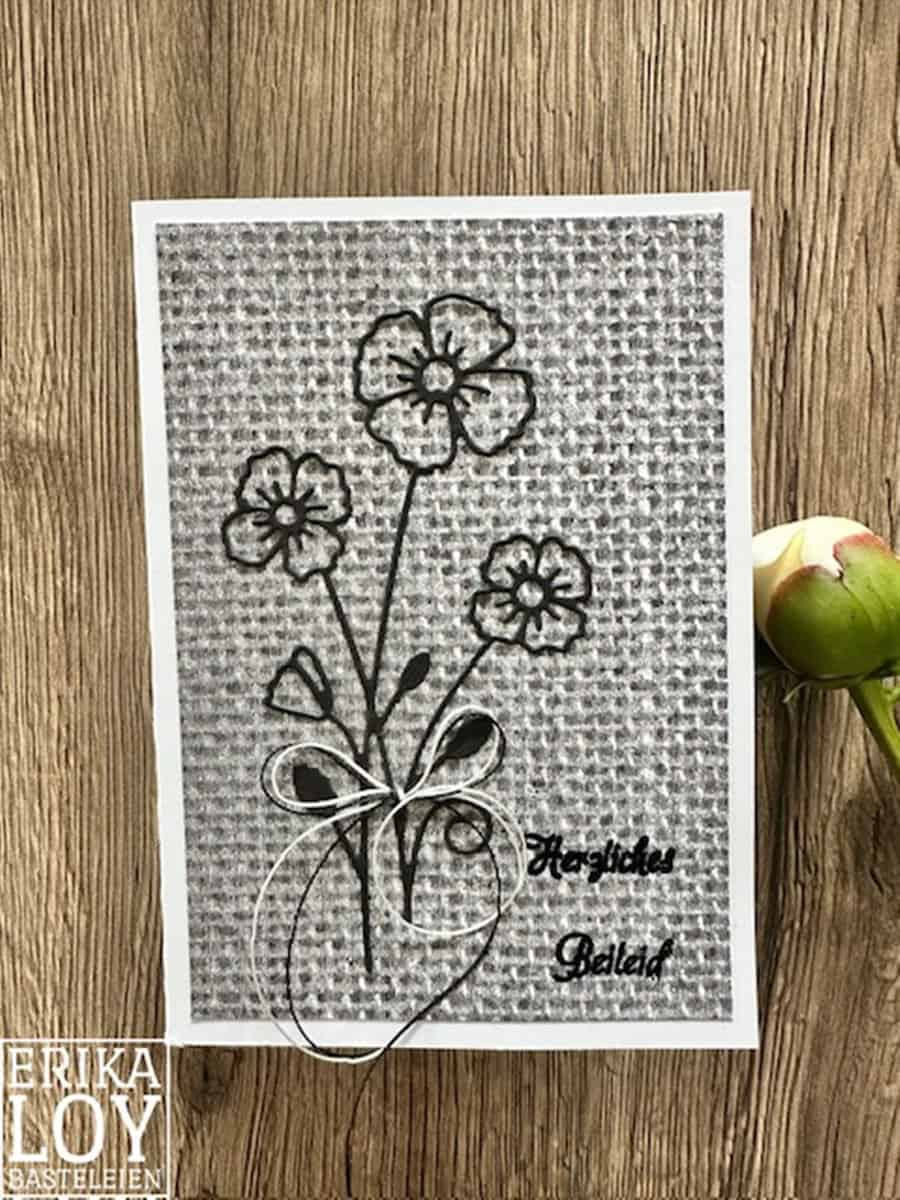Eine handgefertigte Karte mit der Stanzschablone Blume mit drei Blüten von Stanzenshop.de, einer weißen Schleife und "Herzlichen Beileid" auf strukturiertem Grau - ideal zum Basteln, abgebildet auf einer Holzfläche.