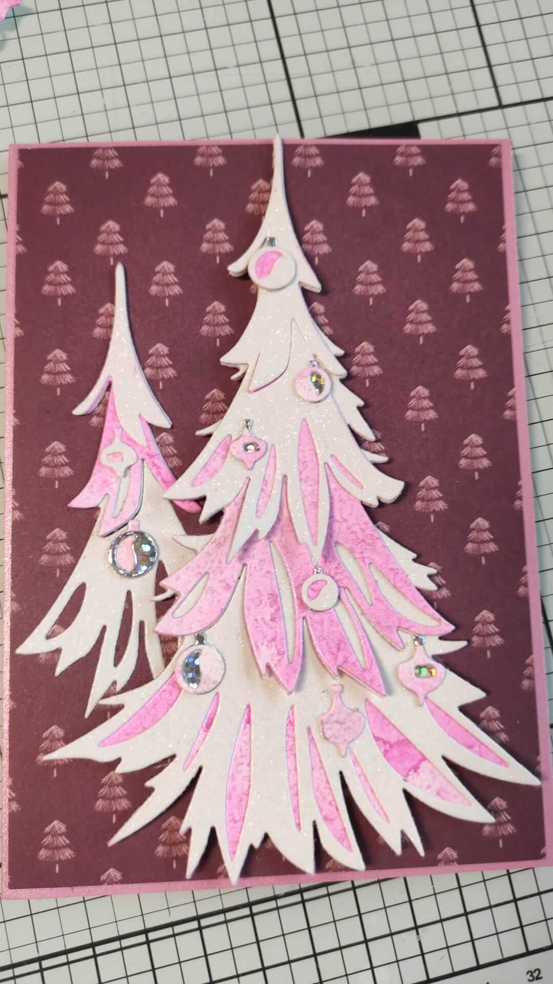 Handgemachte Karte mit geschichteten weißen und rosafarbenen Weihnachtsbäumen und Ornamenten, hergestellt mit den Sizzix 666332 Thinlits Stanzschablonen Trim a Tree Colorize von Sizzix, auf einem lila Hintergrund mit kleinen Baummustern - ideal für Weihnachten.