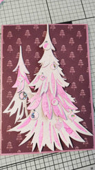 Handgemachte Karte mit geschichteten weißen und rosafarbenen Weihnachtsbäumen und Ornamenten, hergestellt mit den Sizzix 666332 Thinlits Stanzschablonen Trim a Tree Colorize von Sizzix, auf einem lila Hintergrund mit kleinen Baummustern - ideal für Weihnachten.