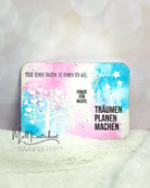 Eine lebendige Karte mit einer weißen Baumsilhouette aus der Stanzschablone: Baum mit Herzen von Stanzenshop.de, mit Sternausschnitten und deutschem Motivationstext auf blau-rosa Aquarellhintergrund - ideal für Scrapbooking oder Bastelzubehör.