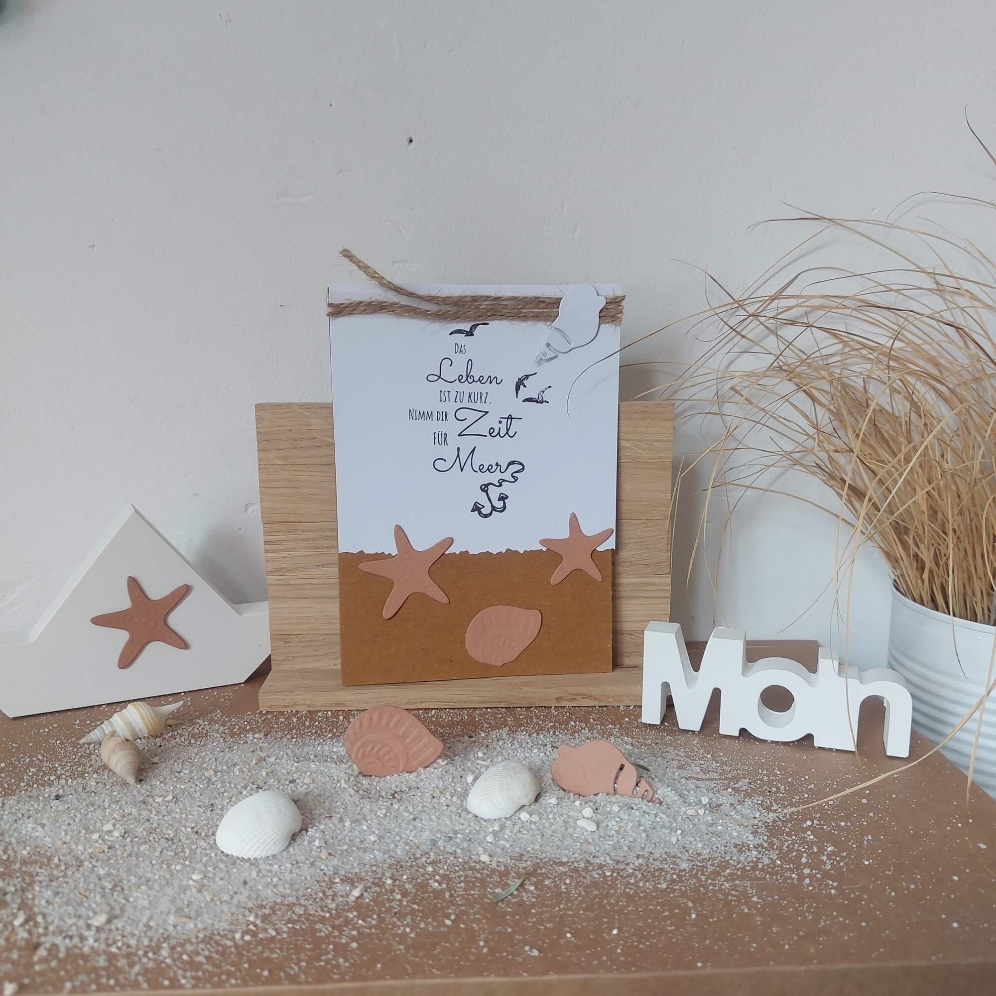 Mit der "Stanzschablone Zwei Muscheln und zwei Seesterne" von Stanzenshop.de lassen sich Muschel- und Seesternformen, eine deutsche Karte und ein weißes "Moin"-Schild auf sandigem Holz perfekt für das nächste Bastelprojekt in Szene setzen!.