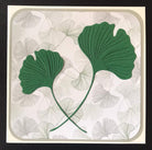 Eine quadratische Karte mit zwei grünen, geprägten Ginkgoblättern aus der Stanzschablone Zwei Ginkgoblätter von Stanzenshop.de auf einem hellen, mit hellgrauen Ginkgo-Konturen gemusterten Hintergrund - ideal zum Basteln oder Scrapbooking.