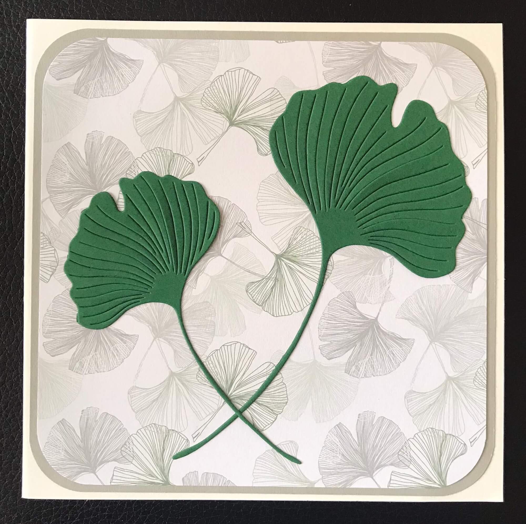 Eine quadratische Karte mit zwei grünen, geprägten Ginkgoblättern aus der Stanzschablone Zwei Ginkgoblätter von Stanzenshop.de auf einem hellen, mit hellgrauen Ginkgo-Konturen gemusterten Hintergrund - ideal zum Basteln oder Scrapbooking.